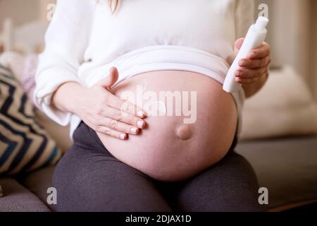 Nahaufnahme der schwangeren Frau, die eine Creme auf ihren Bauch auftragend, während sie zu Hause sitzt. Stockfoto