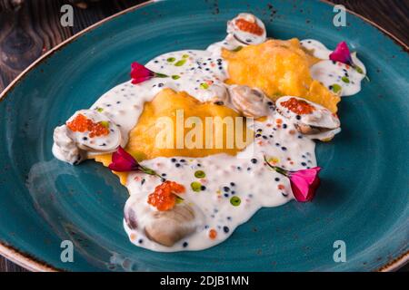 Ravioli mit Krabbe in Kaviarsauce auf einem großen türkisfarbenen Teller. Die Schale steht auf einem Holztisch und ist mit einem leichten Tuch drapiert. Stockfoto