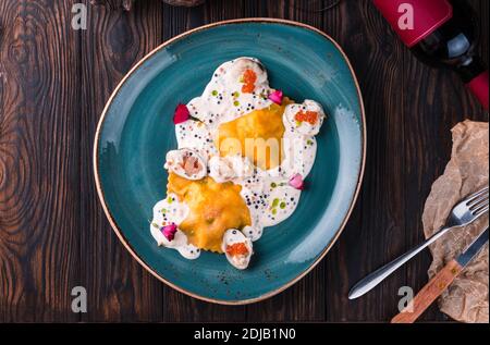 Ravioli mit Krabbe in Kaviarsauce auf einem großen türkisfarbenen Teller. Die Schale steht auf einem Holztisch und ist mit einem leichten Tuch drapiert. Stockfoto