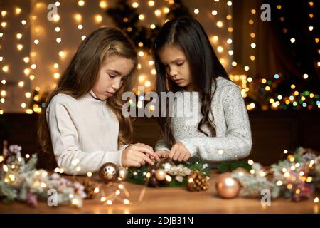 Zwei niedliche kleine Mädchen machen einen handgefertigten Weihnachtskranz zu Hause. Stockfoto