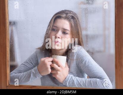 Unglückliche tausendjährige Frau, die Kaffee am Fenster trinkt und den Regen beobachtet, sich melancholisch und einsam fühlt bei schlechtem Wetter Stockfoto