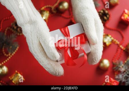 Hände in weißen gestrickten Fäustlingen mit einem Geschenk auf rotem Hintergrund. Weiße Box mit rotem Band. Nachhaltiger Urlaubslebensstil. Weihnachtsschmuck Stockfoto
