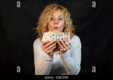Blonde Frau mittleren Alters sieht mit großer Überraschung auf einer Serie aus Von 50 Euro-Banknoten, die sie in den Händen halten Stockfoto