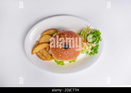 burger auf einem Teller mit Sesamkörnern auf einem weißen Teller. Draufsicht auf Burger und Kartoffelkeile mit Gemüse auf weißem Hintergrund Stockfoto