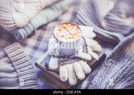 Auf einem Stapel von Büchern und Wollhandschuhen befindet sich ein eleganter Becher Cappuccino mit Marshmallows, daneben liegen warme Winterkleidung aus Wolle: Ein Hut, eine Narbe Stockfoto