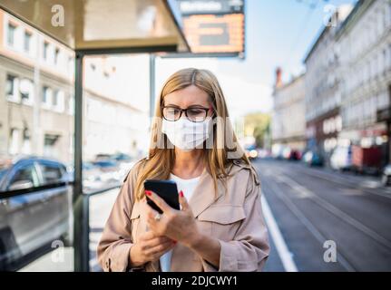 Ältere Frau mit Smartphone an der Bushaltestelle im Freien in der Stadt oder Stadt, Coronavirus Konzept. Stockfoto