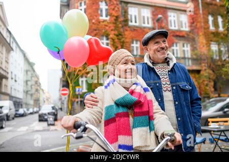 Glückliches Seniorenpaar mit Fahrrad und Luftballons draußen auf der Straße in der Stadt. Stockfoto