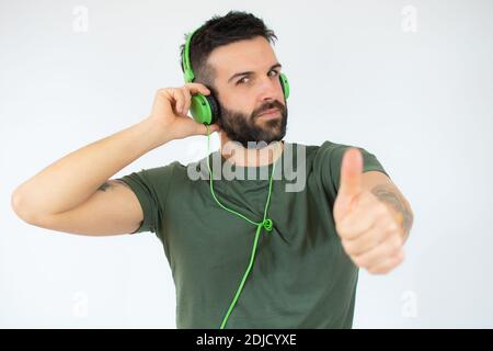 Junger bärtiger Mann mit Kopfhörern auf dem Kopf, der Musik hört, den Daumen nach oben zeigt und isoliert auf weißem Hintergrund mit Kopierraum steht Stockfoto