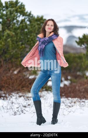 Junge Frau steht im Winterwald in der Nähe immergrüner Nadelbäume und schaut auf die Kamera. Nette Brünette Frau mit langen Haaren, in beige Jacke gekleidet Stockfoto