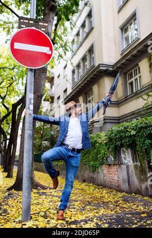 Junger Mann mit Regenschirm, der draußen auf der Straße an einem Lampenpfosten festhält, tik tok Konzept. Stockfoto