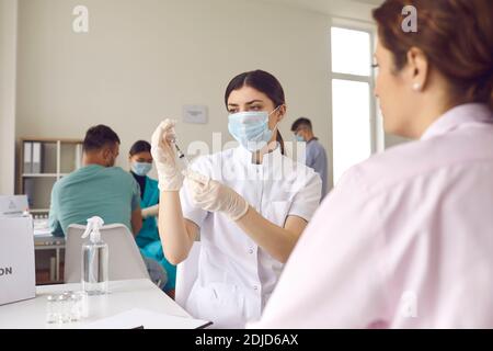 Krankenschwester in einer Maske wählt den Impfstoff in eine Spritze, während sie in einem Krankenhaus neben einem Patienten sitzt. Stockfoto