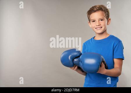 Fröhliche männliche Kind in blauem Hemd Blick auf Kamera und Lächelnd, während Sie Boxhandschuhe halten Stockfoto