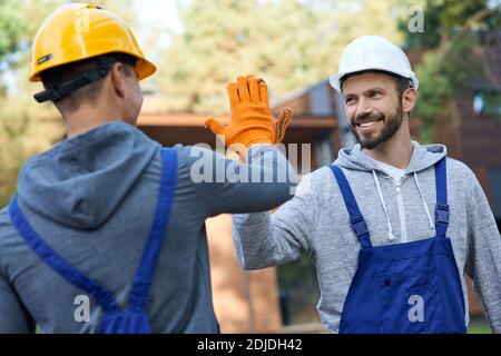 Zwei positive junge Ingenieure in Hartmützen lächeln einander an, geben hoch fünf während der Arbeit auf Hütte Baustelle im Freien. Aufbau, Teamarbeit, Partnerschaftskonzept Stockfoto