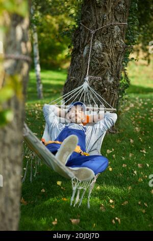 Friedlicher junger Arbeiter, der Uniform trägt, eine Pause macht, in einer Hängematte draußen liegend, mit geschlossenen Augen an einem sonnigen Tag. Gebäude, Beruf, Rest-Konzept Stockfoto