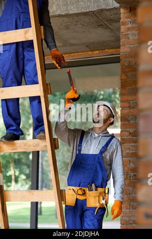 Junger Arbeiter in Overalls und Harthut, der seinem Kollegen auf der Leiter Hammer gab, während er auf der Hütte-Baustelle arbeitete. Building, People, Teamwork-Konzept Stockfoto