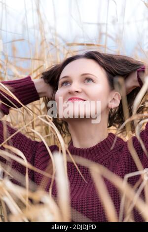 Porträt von stilvollen jungen Frau in braunen Pullover posiert in getrocknetem Gras im Feld gekleidet. Glückliche Frau mit langen lockigen Brünette Haare und roten Lippen. Stockfoto