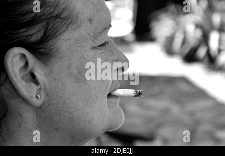 Alte Frau mit Flecken im Gesicht und Warze, Rauchen eine Zigarette in der Tür ihres Hauses, Seitenansicht, schwarz-weiß Foto. Stockfoto