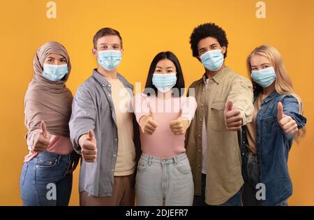 Internationaler multiethnischer Studentenaustausch. Junge multirassische Menschen in Schutzmasken zeigen Daumen nach oben Stockfoto