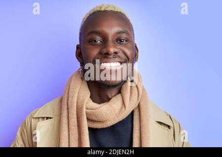 Optimistisch lächelnder Afro Kerl in trendigen Mantel Outfit, lächeln an der Kamera mit perfekten toothy Lächeln, Mode-Modell isoliert über lila Hintergrund Stockfoto