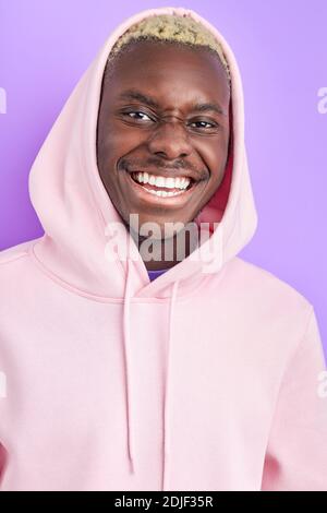 Optimistisch lächelnd Afro Kerl in trendigen rosa Pullover, lächeln an der Kamera mit perfekten toothy Lächeln, Mode-Modell isoliert über lila Hintergrund Stockfoto