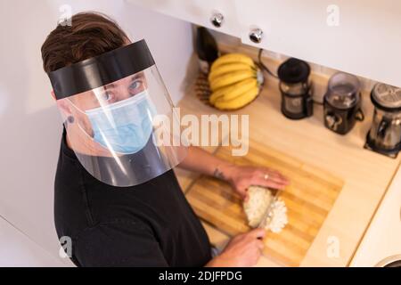 Nahaufnahme von oben von einem jungen Mann in der Küche, hacken eine Zwiebel auf einem schneidenden Wildschwein tragen covid Schutz Gesichtsmaske und Kunststoff-Visier Stockfoto