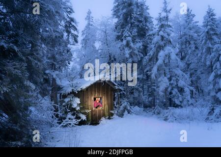 Frau, die aus dem Fenster eines einsamen Holzhauses in einem tief verschneiten Tannenwald in den Allgäuer Alpen schaut Stockfoto