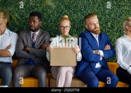 Interracial Team von Mitarbeitern unglücklich mit der Ankunft neue Emplyee, blonde kaukasische Frau in Brillen sitzt mit Box von Dingen, Blick auf die Kamera, traurig Stockfoto