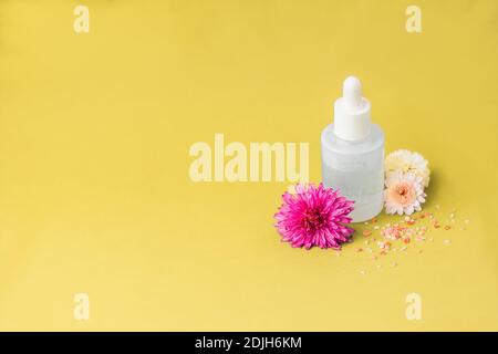 Beauty-Produkt-Paket auf gelbem Hintergrund, rosa Blumen und Meersalz bestreut. Leere weiße Serumflasche mit Pipette und ohne Branding. Text platzieren Stockfoto