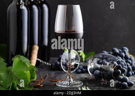 Weinglas mit kaltem Rotwein. Weinflaschen, Weintrauben mit Blättern und Reben auf dunklem rustikalem Betonhintergrund. Weinkomposition auf schwarzem Stein Stockfoto