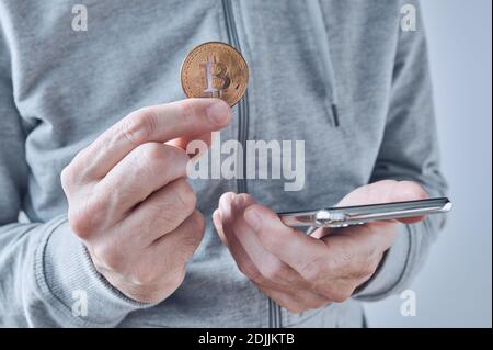 Bitcoin Trading App für Smartphones, konzeptionelles Bild, Mann hält Kryptowährung Münze und Handy in Händen. Stockfoto