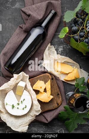 Rotwein Flasche Camembert Käse und gereiften Käse, Trauben. Snacks an der Weinbar. Weinkomposition auf dunklem rustikalem Betonhintergrund. Flach auflagertes vertikales Erntegut Stockfoto