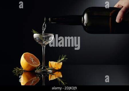 Gin wird in ein kleines Glas aus einer antiken Flasche aus dunklem Glas gegossen. Ein starkes alkoholisches Getränk mit Zitrone und Rosmarin auf einem schwarzen reflektierenden Hintergrund Stockfoto
