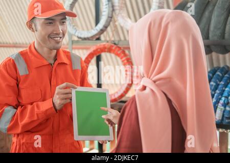 Mechaniker in einem Wearpack trägt ein digitales Tablet mit einem Kunde im Hintergrund eines Reifenregals Stockfoto