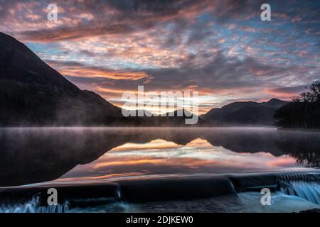 Ein schöner Wintersonnenaufgang am Wehr am Crummock Water, mit Nebel und einem goldenen Schein auf Wolken, die sich im See spiegeln. Lake District, Cumbria, England Stockfoto