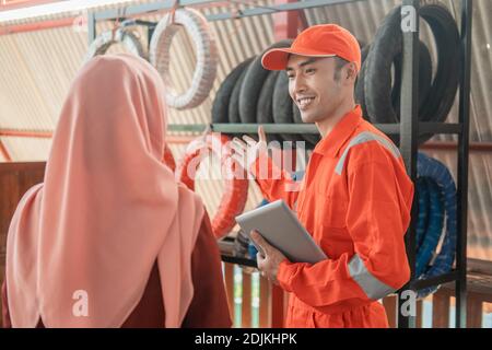 Mann Mechaniker in Wearpack Uniform hält digitale Tablette während des Serviers Verschleierte weibliche Kunden, die in der Werkstatt Reifen auswählen Stockfoto