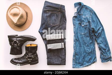 Herren Mode-Outfit Gitter. Einfacher weißer Hintergrund. Jeans aus Denim, Jeanshemd mit Perlmutt-Druckknöpfen, brauner Hut, schwarze Stiefel. Stockfoto