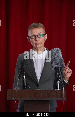 Vertikales Porträt der reifen Frau, die Rede auf dem Podium stehend Auf der Bühne gegen roten Vorhang Stockfoto