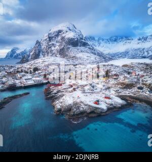 Landschaft mit blauem Meer, schneebedeckten Bergen, hohen Felsen, Gebäuden, traditionellen Rorbu, bewölktem Himmel. Luftaufnahme des kleinen Dorfes bei Sonnenuntergang im Winter. Reise Stockfoto