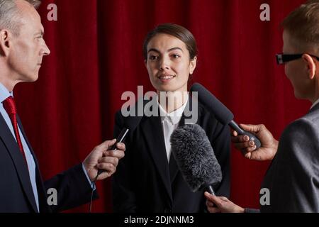 Waist up Porträt der jungen Frau geben Interview Journalist Und mit Mikrofonen gegen roten Vorhang sprechen Stockfoto