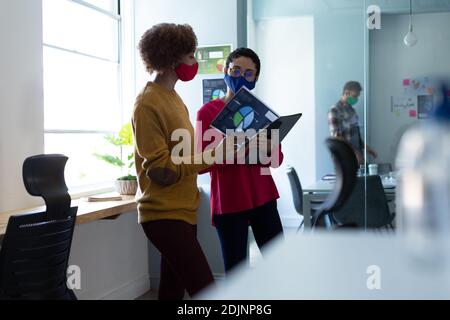 Zwei Frauen mit gemischter Rasse, die Masken tragen und in einem Büro sprechen Stockfoto
