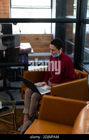 Mixed Race Frau trägt gesenkt Gesichtsmaske mit Laptop und Kopfhörer für Videokonferenzen im Büro Stockfoto