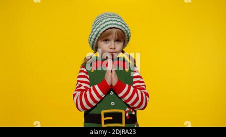 Child girl pleads about gifts on New Year. Pleading kid in Christmas elf Santa helper costume begs, prays for forgiveness feels sorry and guilty, keeps palms together, standing over yellow background Stock Photo