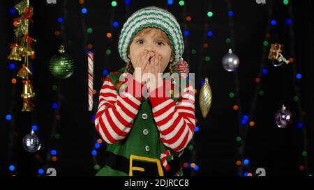 Beeindruckt Kind Kind Mädchen in Weihnachten Elf Santa Claus Helfer Kostüm sieht in der Kamera macht große Augen decken Mund mit Händen fühlen sich entsetzt isoliert auf schwarzem Hintergrund. Oh Mann, wow, fassungslos, schockiert Stockfoto