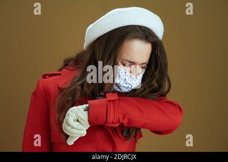 Leben während covid-19 Pandemie. Moderne Hausfrau in rotem Mantel Niesen in Ellbogen auf beigem Hintergrund. Stockfoto