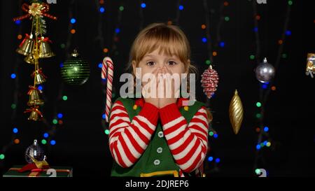 Beeindruckt Kind Kind Mädchen in Weihnachten Elf Santa Claus Helfer Kostüm sieht in der Kamera macht große Augen decken Mund mit Händen fühlen sich entsetzt isoliert auf schwarzem Hintergrund. Oh Mann, wow, fassungslos, schockiert Stockfoto