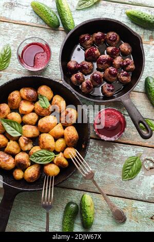 Gesunde nordische Mahlzeit. Bratkartoffeln, Fleischbällchen und Preiselbeergetränk serviert mit rohen Gurken und Basilikum auf rustikalem blauen Tisch oben Stockfoto