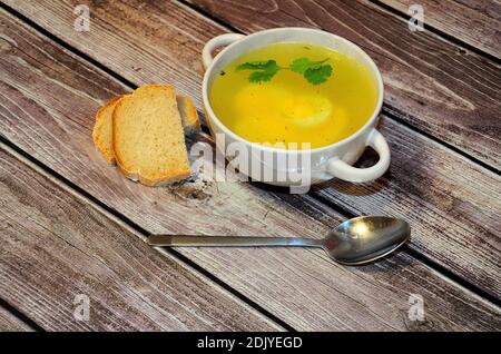 Tiefe Keramikplatte mit frischer Hühnerbrühe, Ei und einem Zweig Koriander auf einem Holztisch, neben einem Löffel und zwei Scheiben Weißbrot. Nahaufnahme. Stockfoto