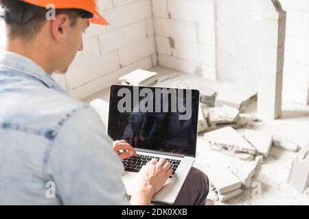 Schöner Architekt oder Vorarbeiter im Helm, der mit Laptop arbeitet Die Wohnung Zeichnungen an der Struktur weiß Interieur Stockfoto