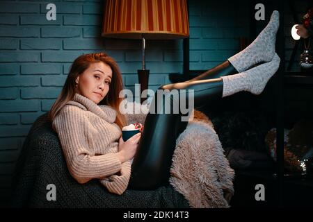 Frau mit Tasse Kakao auf dem Stuhl. Konzept von Weihnachten und Neujahr. Schönes Mädchen in Pullover auf Strickdecke. Graue Ziegelwand auf dem Hintergrund. Winterurlaub. Gestreifte Stehleuchte hinter dem Stuhl. Stockfoto