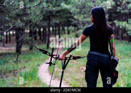 A back view of a woman hunter holding a compound bow and arrow in a forest Stock Photo